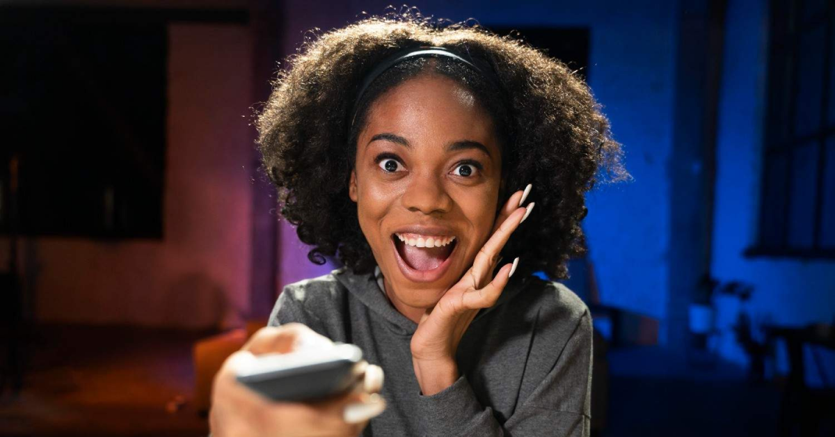 Mulher sorridente assiste TV com controle remoto na mão, expressando empolgação.