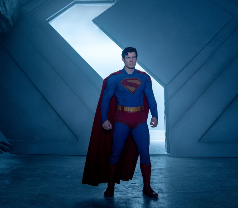 David Corenswet caracterizado como Superman no filme de 2025, usando traje azul, capa vermelha e cinto dourado, em um cenário futurista.